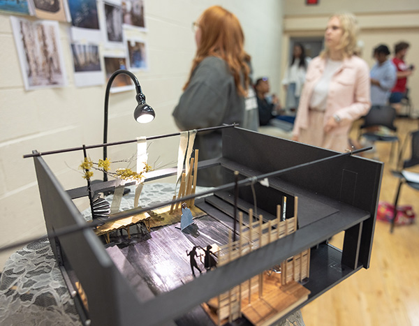 Close-up of a small foam set model, on which are visible a partial lathe wall, a spiral staircase, and strips of fabric hanging from a bar. The set features several two-dimensional human figures, and is lit by a small goose-neck lamp. Out of focus in the background are several people looking at images hung on the wall and conversing.